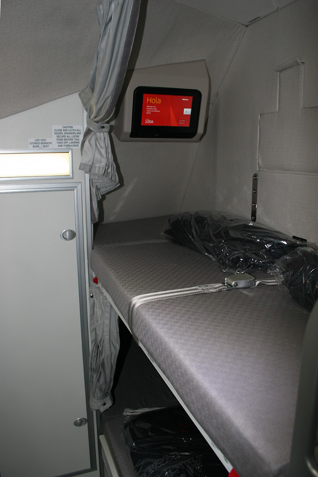 A narrow crew rest bunk with a curtain, a small screen and storage inside an Iberia Airbus A330
