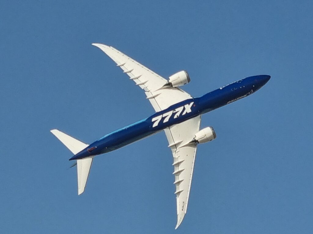 Boeing 777-9 test aircraft in blue and white 777X livery banking in flight, seen from below