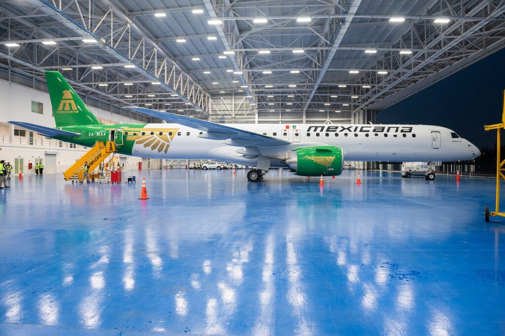 A white and green Mexicana Embraer E195-E2 parked inside a brightly lit hangar with stairs at the rear door