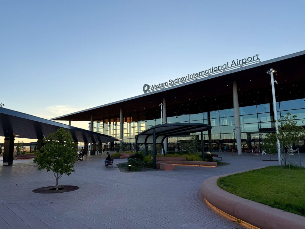 The forecourt and glass terminal of Western Sydney International Airport at dusk, with a few people walking outside
